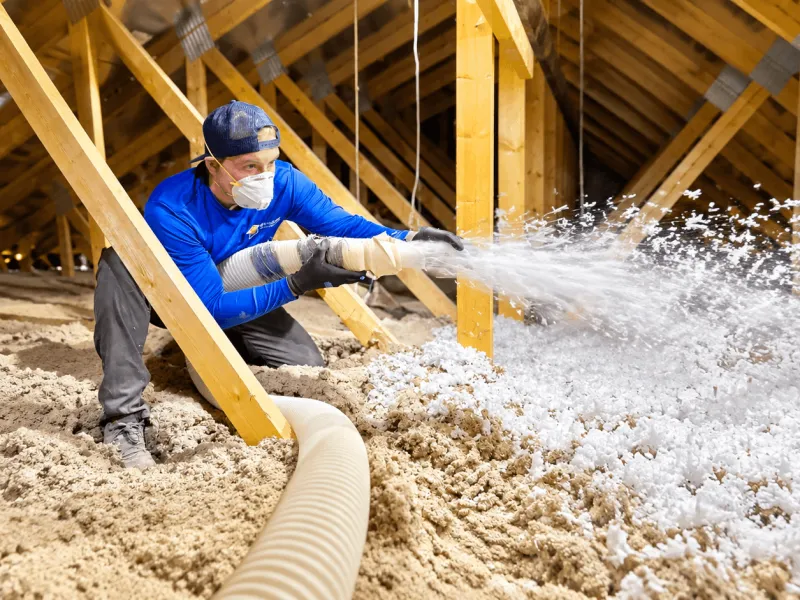 Installer in a dust mask blowing loose-fill insulation through a hose across a timber-framed roof cavity